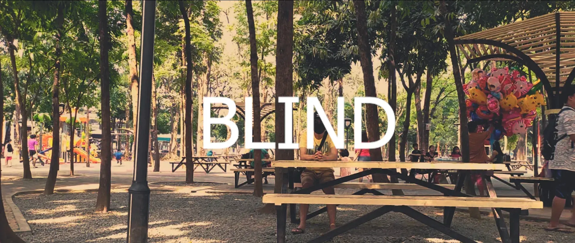 Title card for the short film 'Blind', showing a sunlit park with picnic tables under tall trees