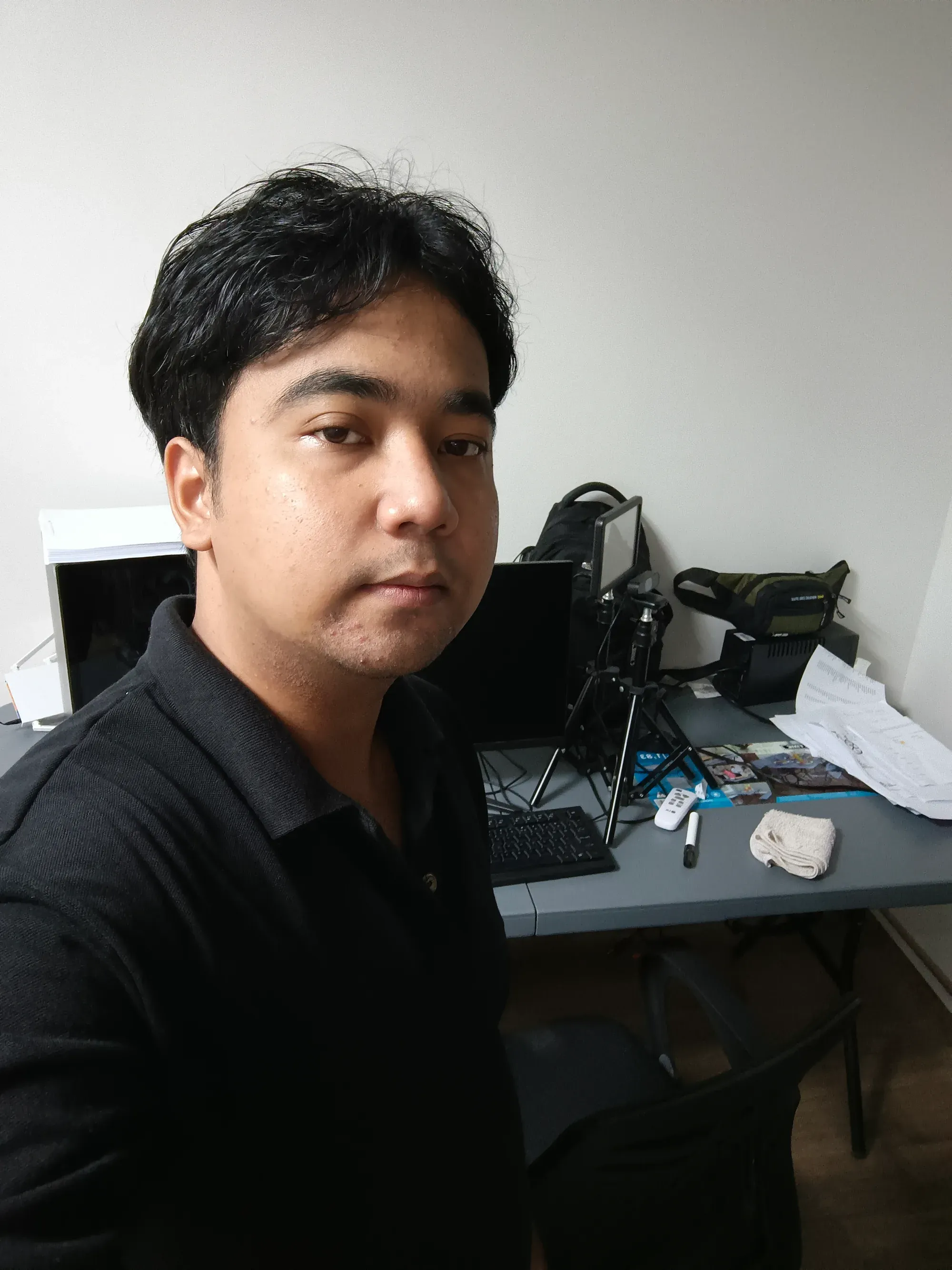 Jon Arbell in a black polo at his desk, a monitor, camera tripod and papers behind him.