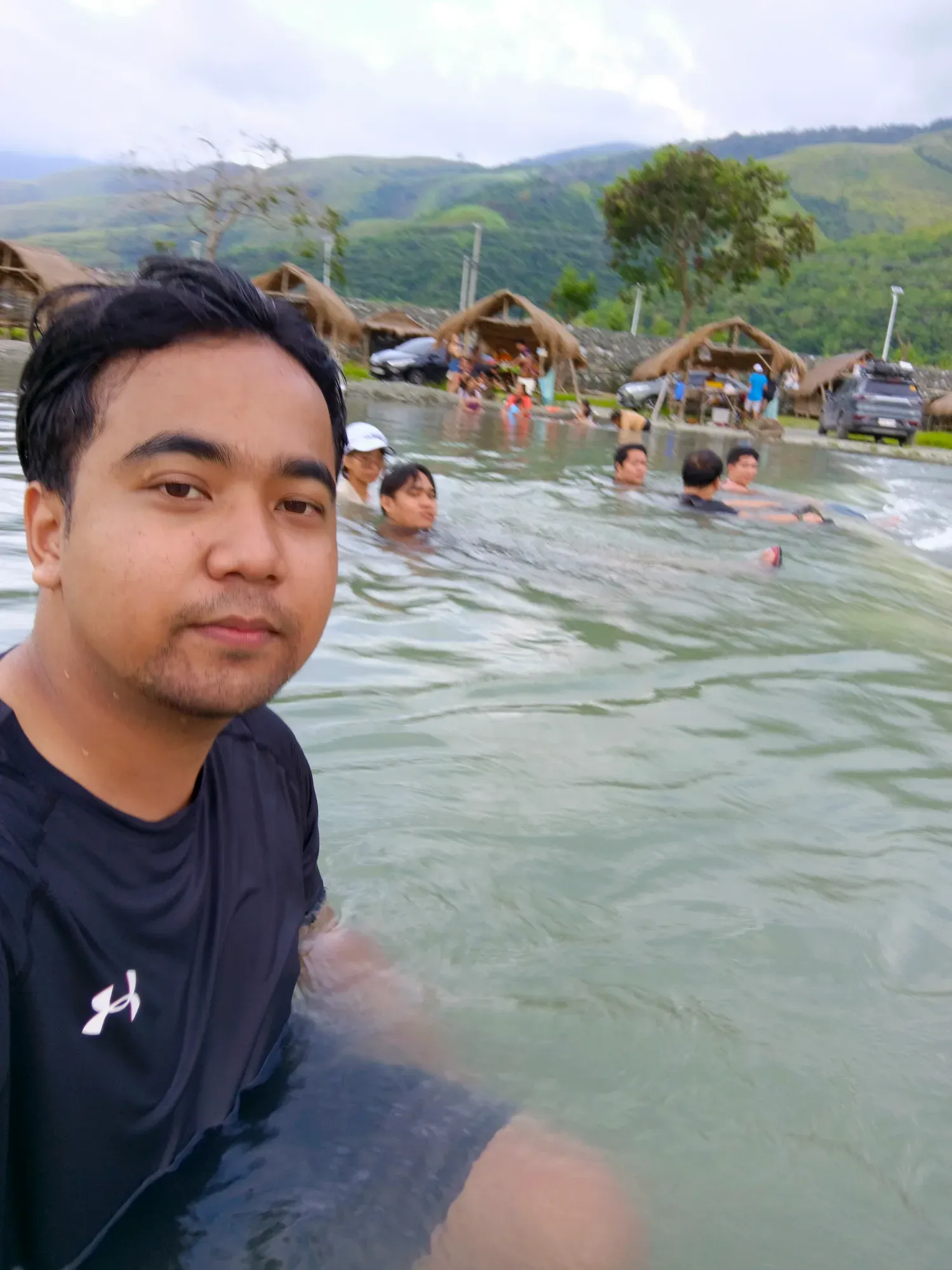 Jon Arbell chest-deep in a mountain hot spring, nipa huts and green ridges behind him.