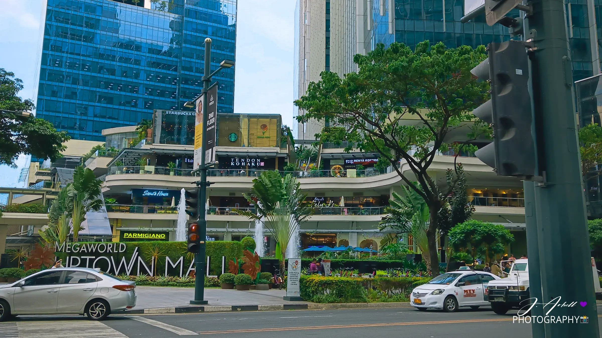 The Uptown Mall facade in BGC on a bright day, traffic passing beneath a clear blue sky