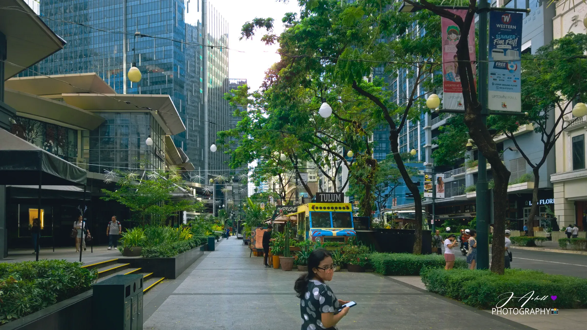 A tree-lined pedestrian promenade in Bonifacio Global City, a lone figure walking between glass office towers