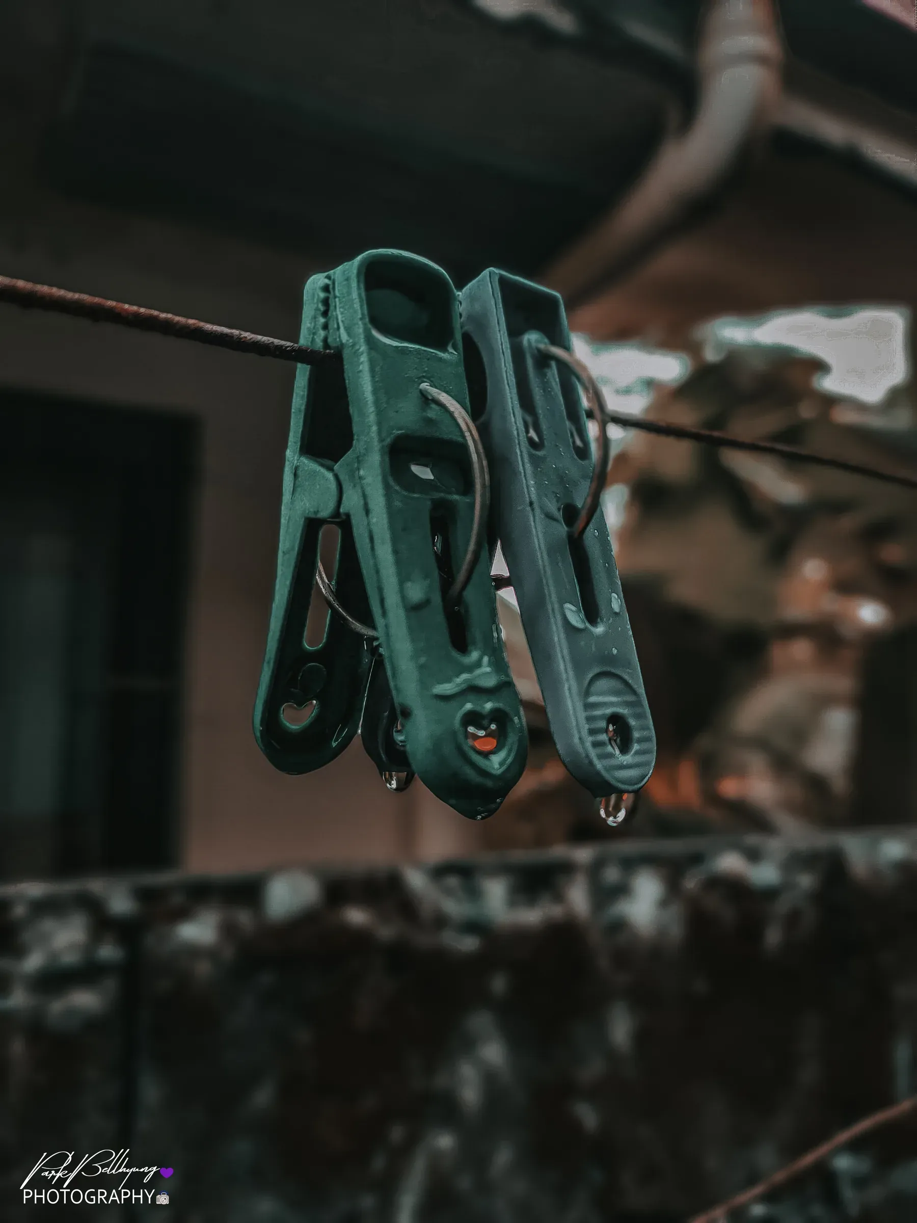 Two green plastic clothespins clipped to a wire with water beading on them, a house wall and eaves blurred out behind