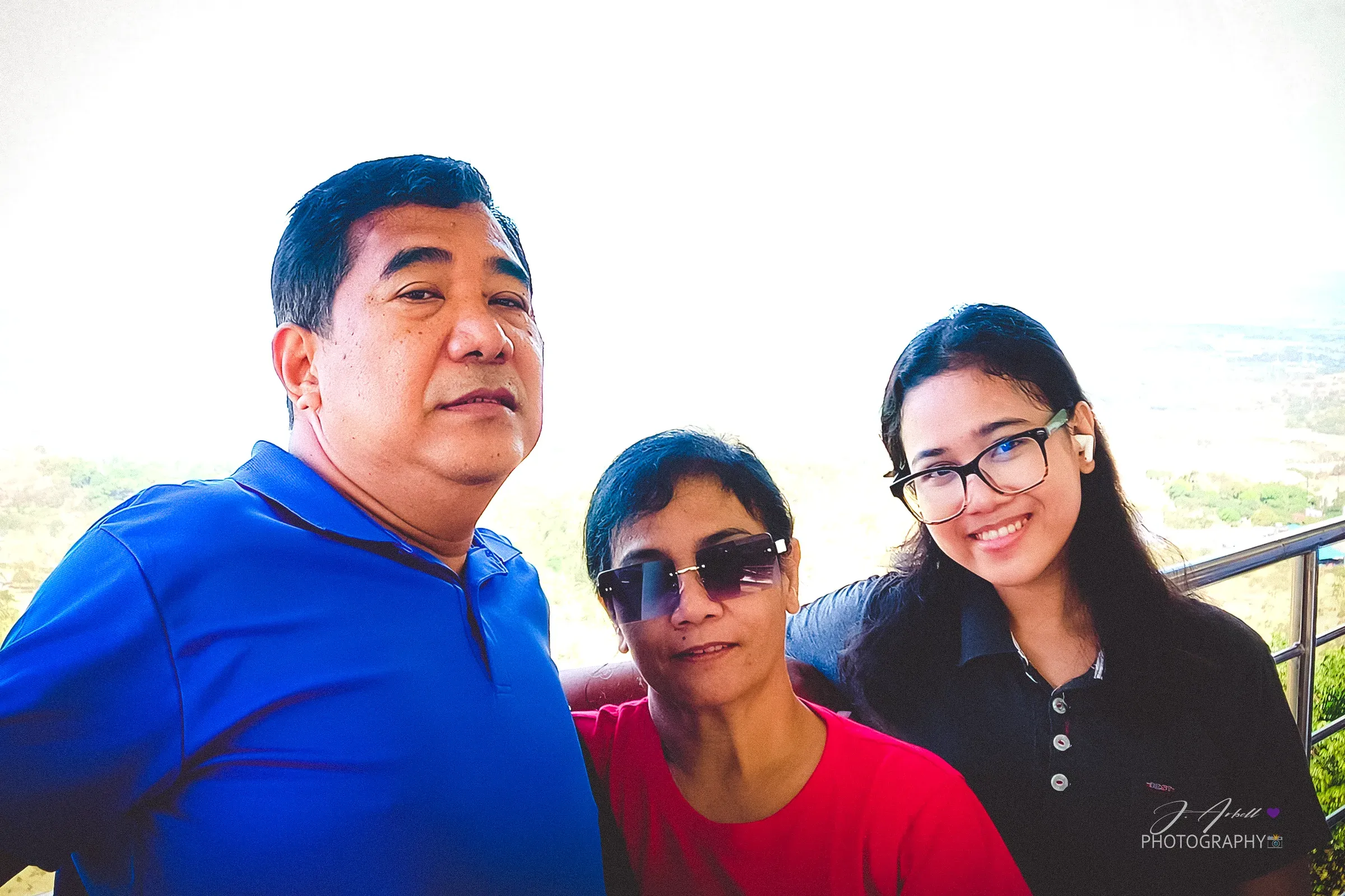 Three family members close together on a viewing deck — a man in a blue polo, a woman in red with sunglasses, and a young woman in glasses — a hazy valley behind them