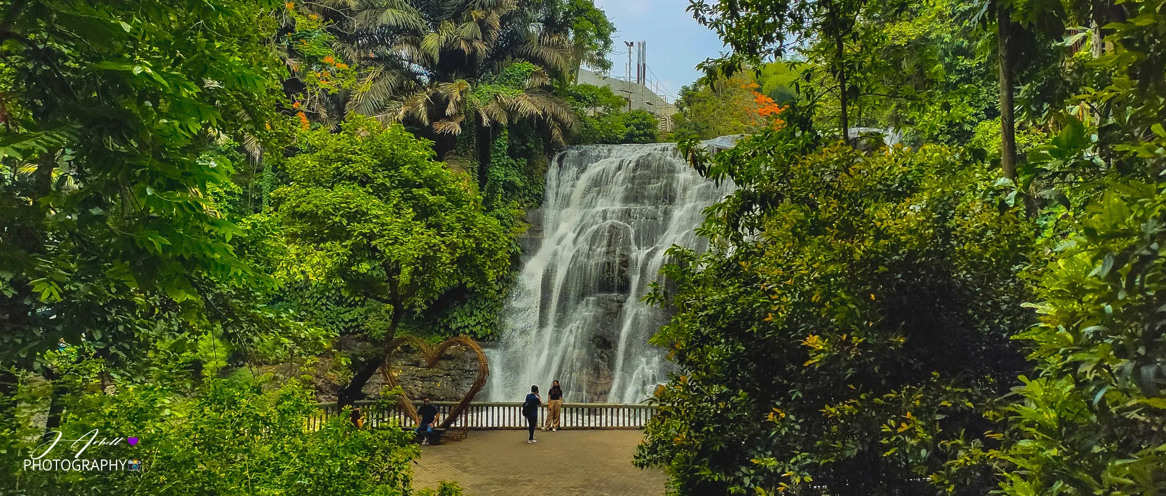 Hinulugang Taktak waterfall in Antipolo framed by dense green foliage, a woven heart sculpture and a few visitors at the railing below the drop