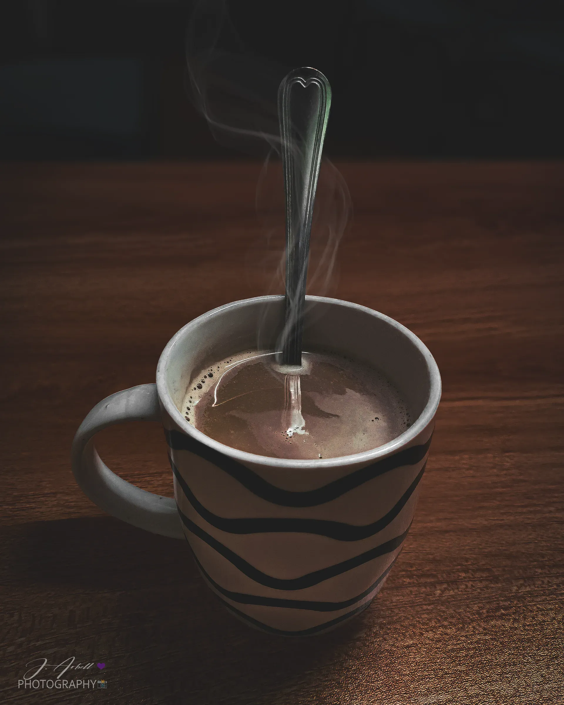 A steaming mug of hot chocolate with a spoon resting in it, on a dark wooden table