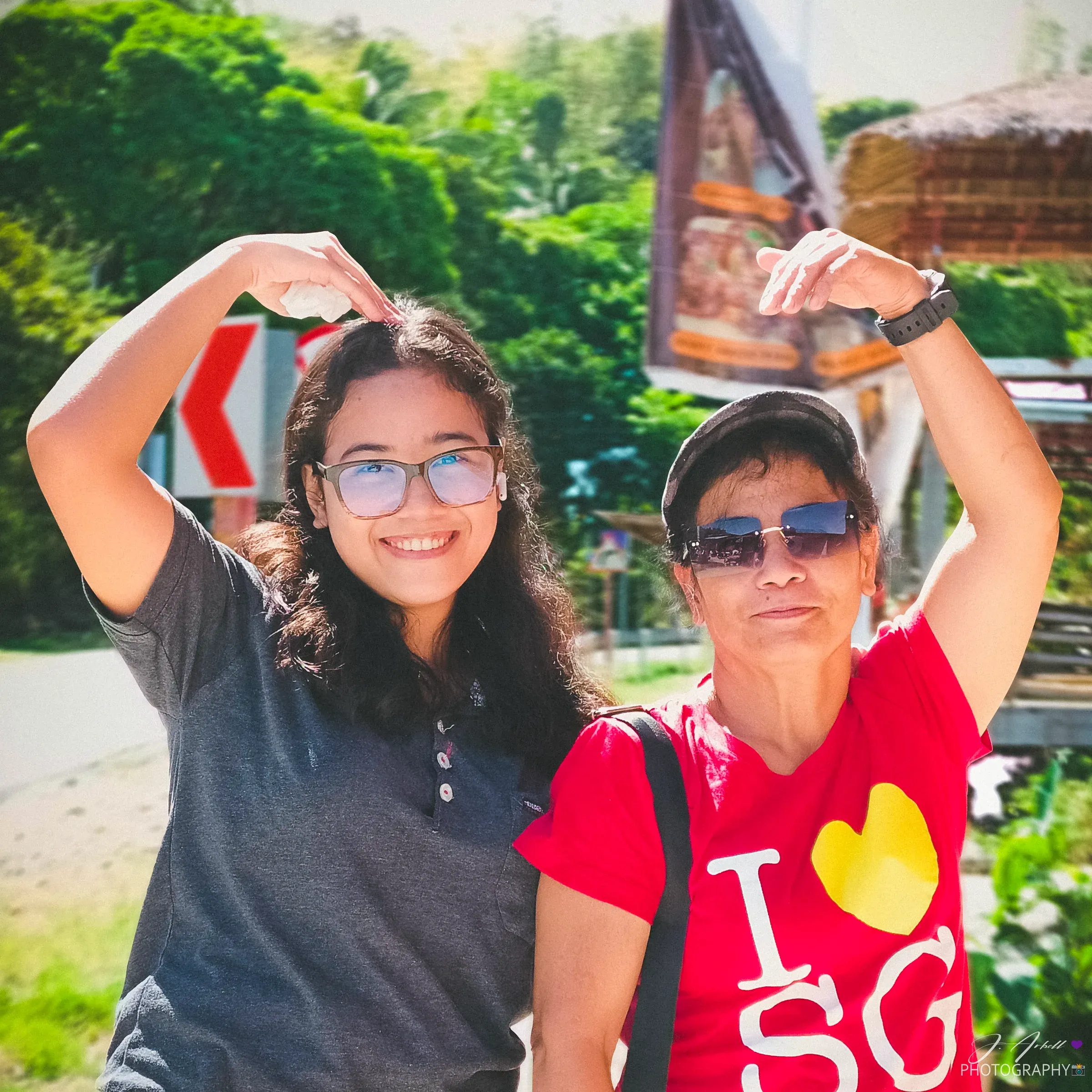Two family members making a heart shape with their arms above their heads at a roadside stop, roadside greenery and a nipa hut behind them