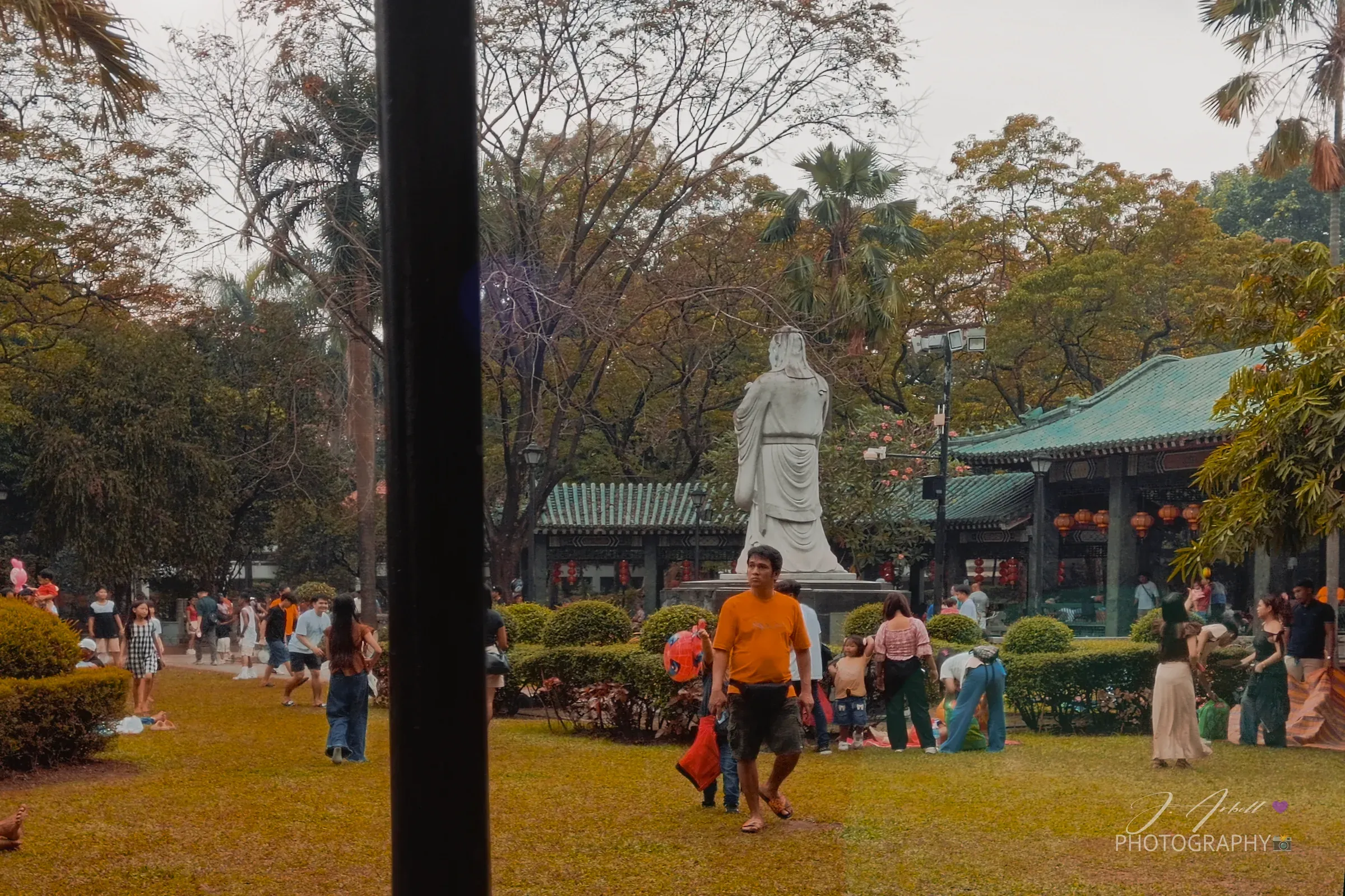 The Chinese Garden at Luneta seen past a dark lamp post, a white robed statue on a plinth in front of green-tiled pavilions hung with red lanterns, visitors spread across the lawn