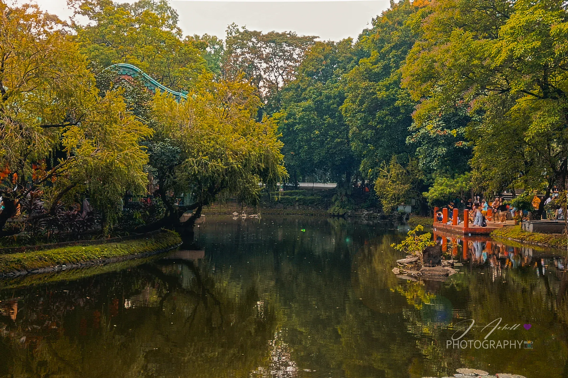 The lagoon at Luneta Park, autumn-toned trees mirrored in still water with visitors gathered along the far bank