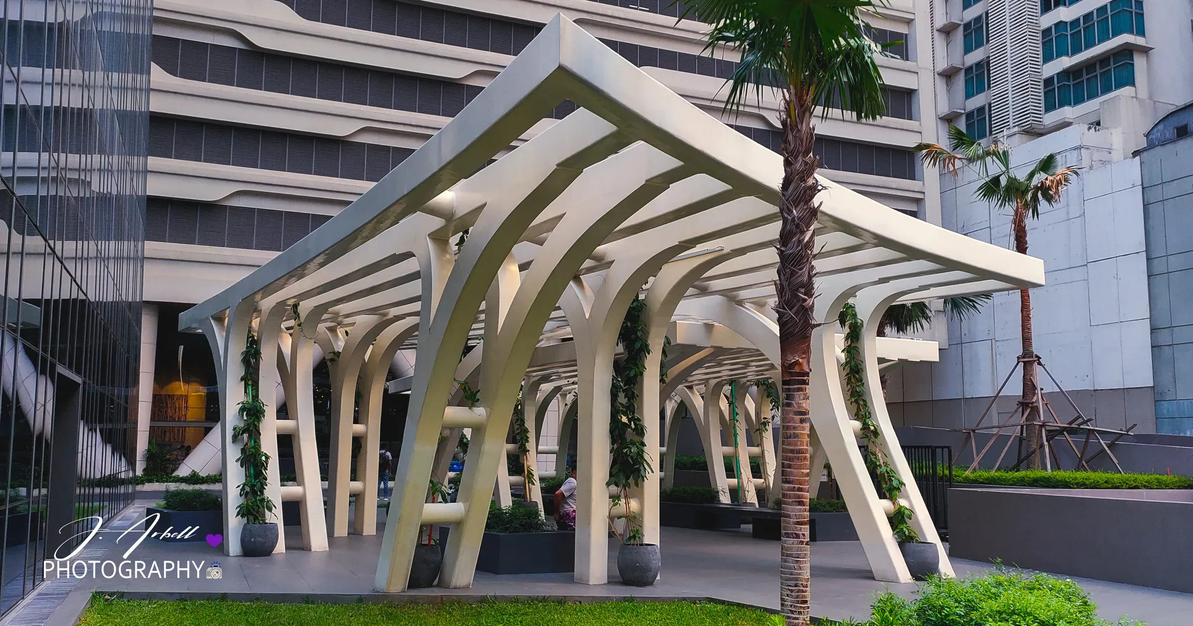 A white arched canopy pavilion beside a palm tree, framed by office towers in Poblacion, Makati