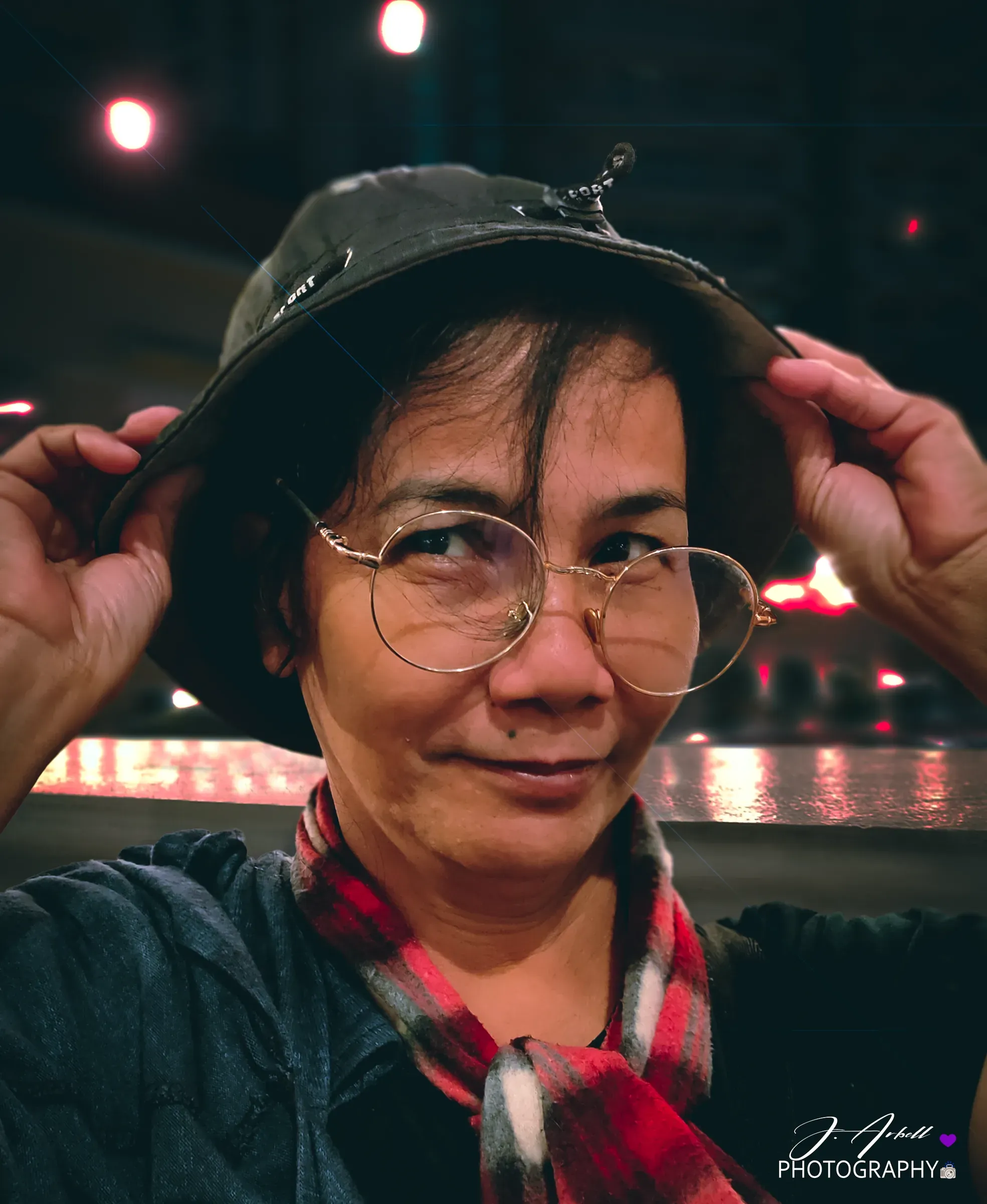 A woman in round glasses and a bucket hat holding the brim with both hands, a red plaid scarf at her neck, warm night lights reflecting on water behind her