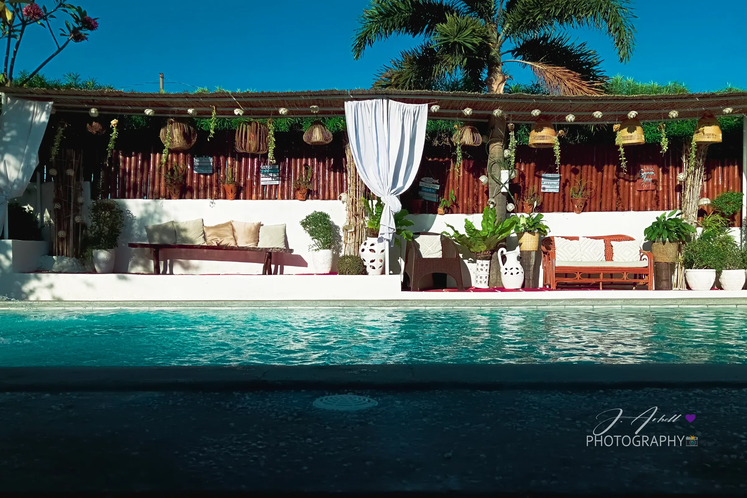A poolside lounge in Calatagan at midday, bamboo screens and cushioned benches reflected in turquoise water