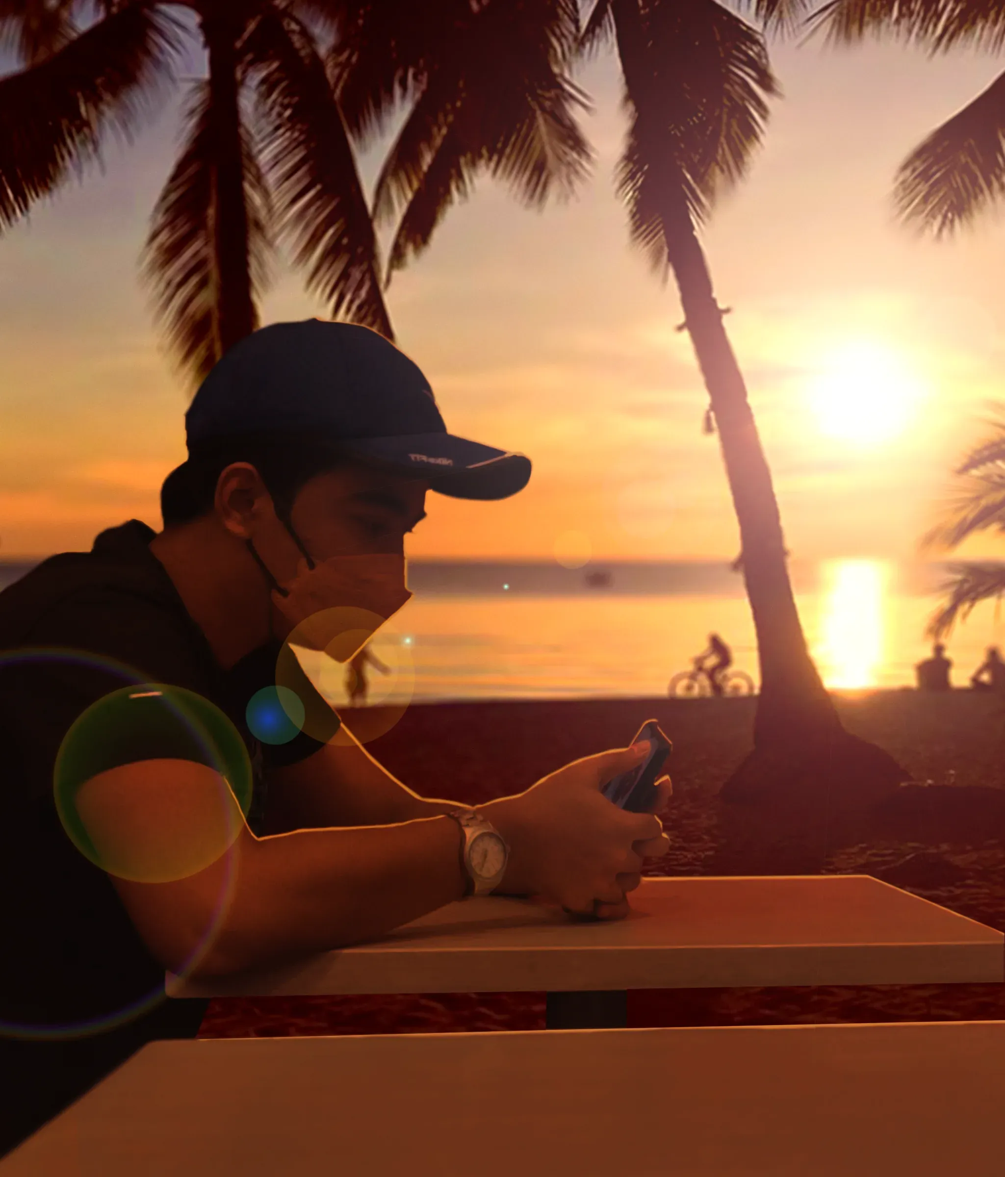 A young man in a cap and face mask at a beachside table looking at his phone, silhouetted against a sunset over the sea with palms and lens flare across the frame