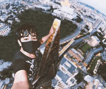 Composite of a masked figure taking a selfie from the very top of a skyscraper, the city laid out far below