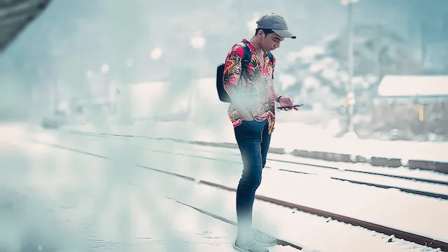 A figure in a bright patterned jacket standing on snow-covered railway tracks, checking a phone
