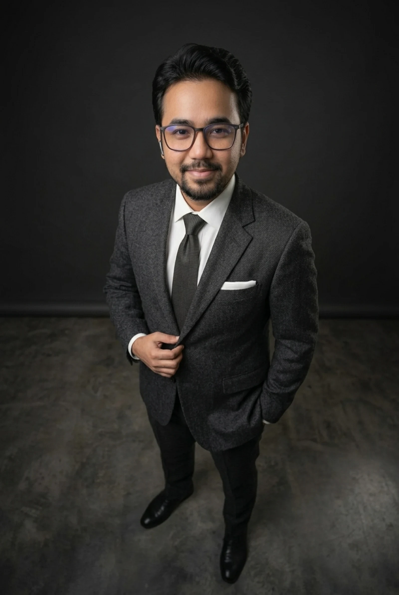 Jon Arbell De Ocampo in a charcoal suit, tie and glasses, photographed from above standing against a dark studio backdrop.