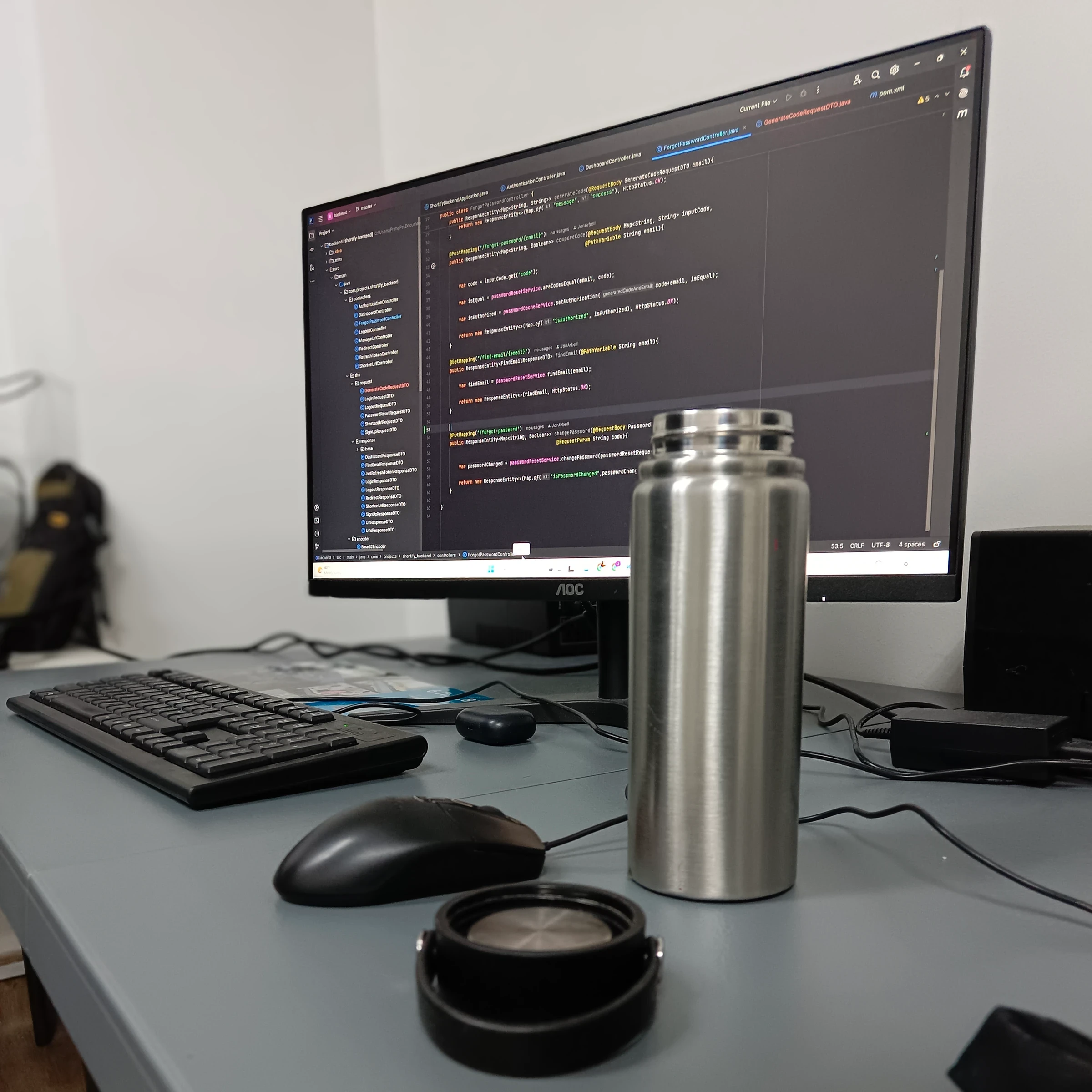 A desk with an external monitor showing a Java Spring Boot controller open in an IDE, project tree beside it, with a keyboard, mouse, water bottle and headphones on the desktop.